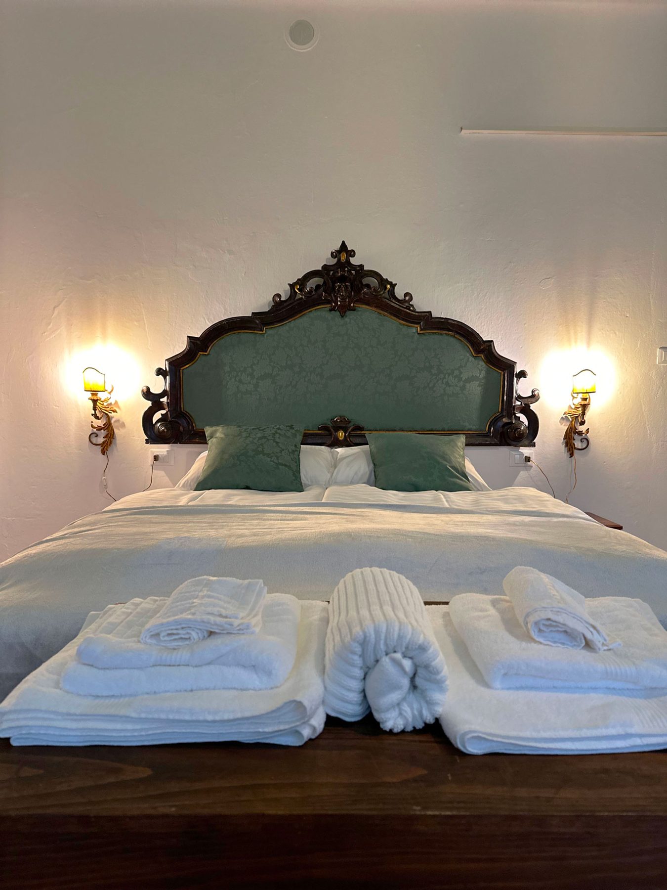 Un letto con la testiera intagliata e verde, gli asciugamani piegati sulle coperte e due lampade accese ai lati